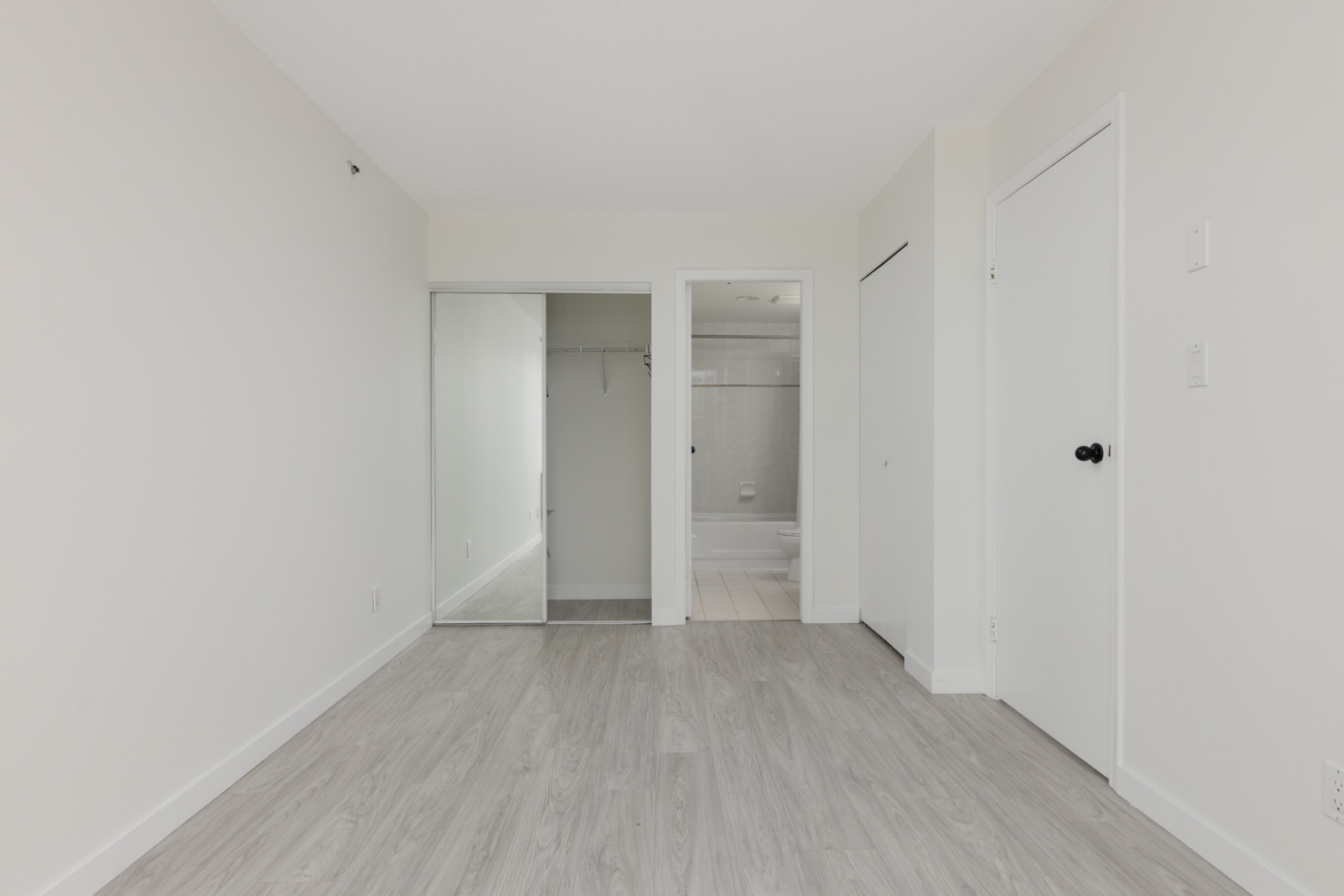Empty room with light wood floors, white walls, mirrored closet, bathroom door, and another door at 950 Cambie St, Vancouver.