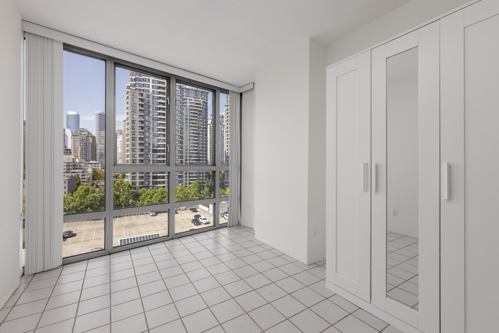 Empty room with tiled floor, large windows facing city buildings, and a white mirrored wardrobe on the right at 950 Cambie St, Vancouver.