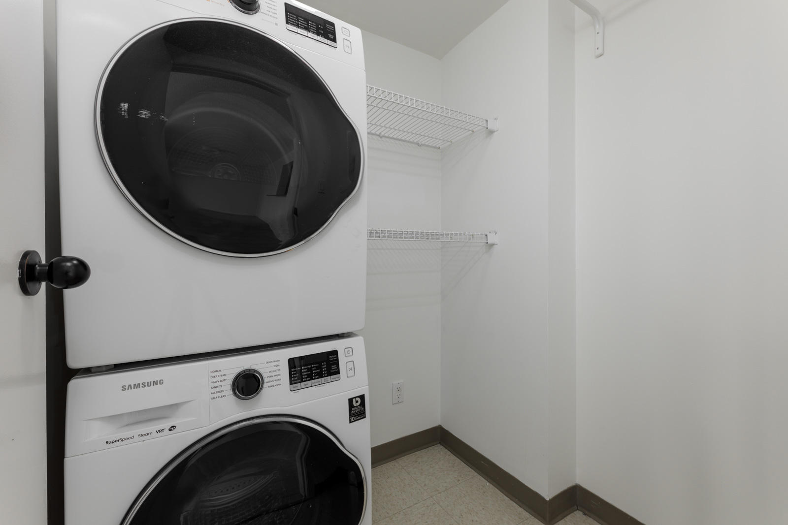 Stacked washer and dryer beside empty white wire shelves in a compact white laundry room with beige trim and tiled floor in Vancouver.