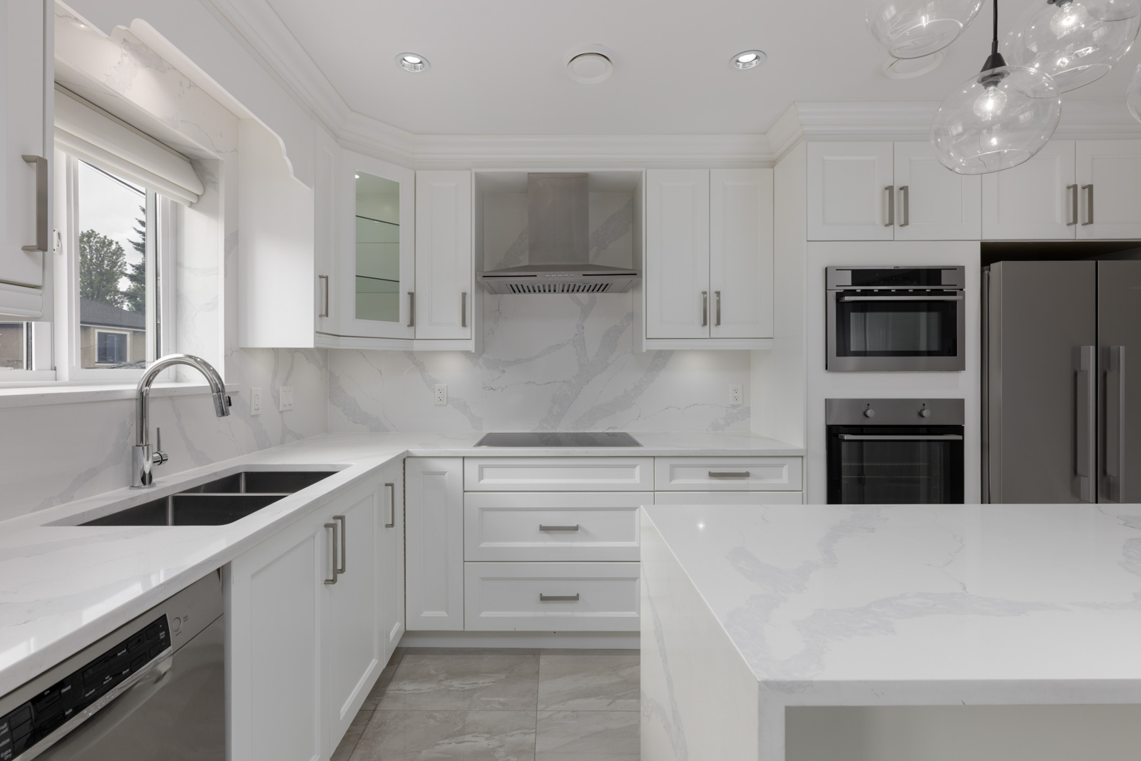 Bright white kitchen with marble counters, stainless appliances, island, double sink, and large window in a Vancouver home on Falaise Ave.