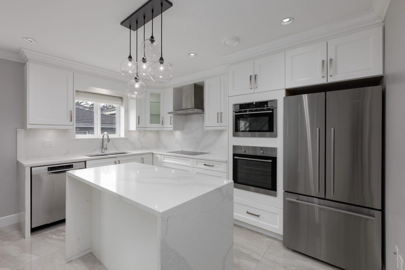 Modern kitchen with white cabinets, marble counters, stainless appliances, island, pendant lights, and large windows in Falaise Place, Vancouver.