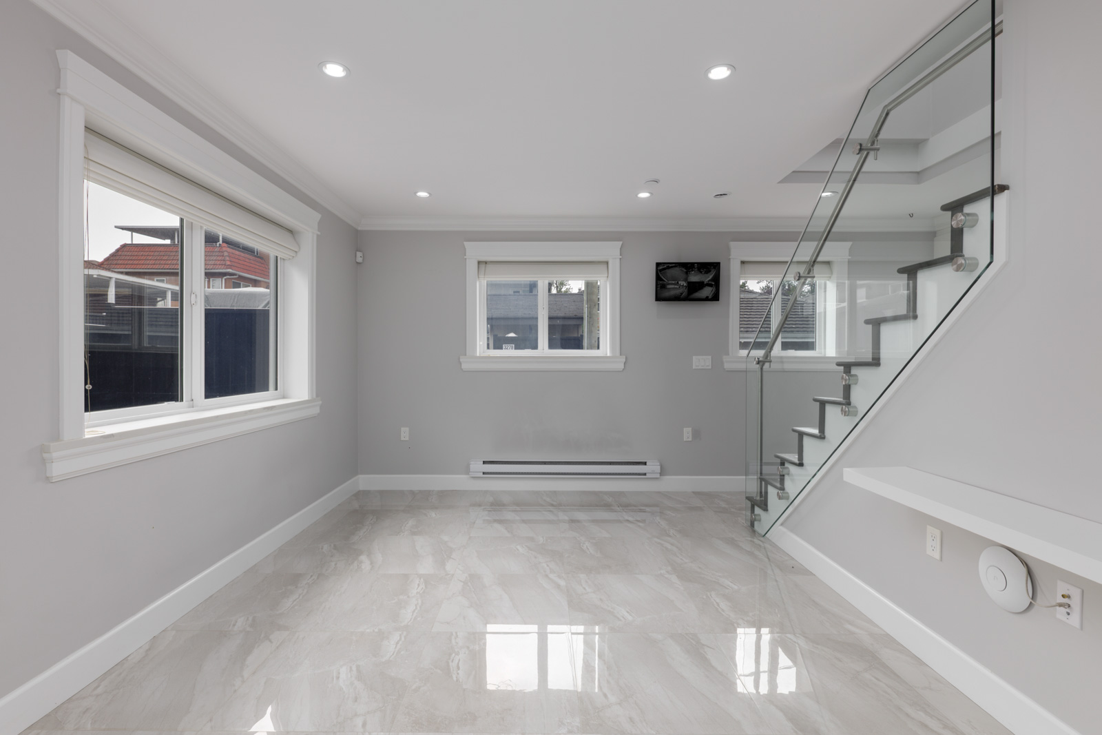 Empty modern room with gray walls, large windows, glossy tile floors, recessed lighting, and glass-railed stairs in Vancouver.
