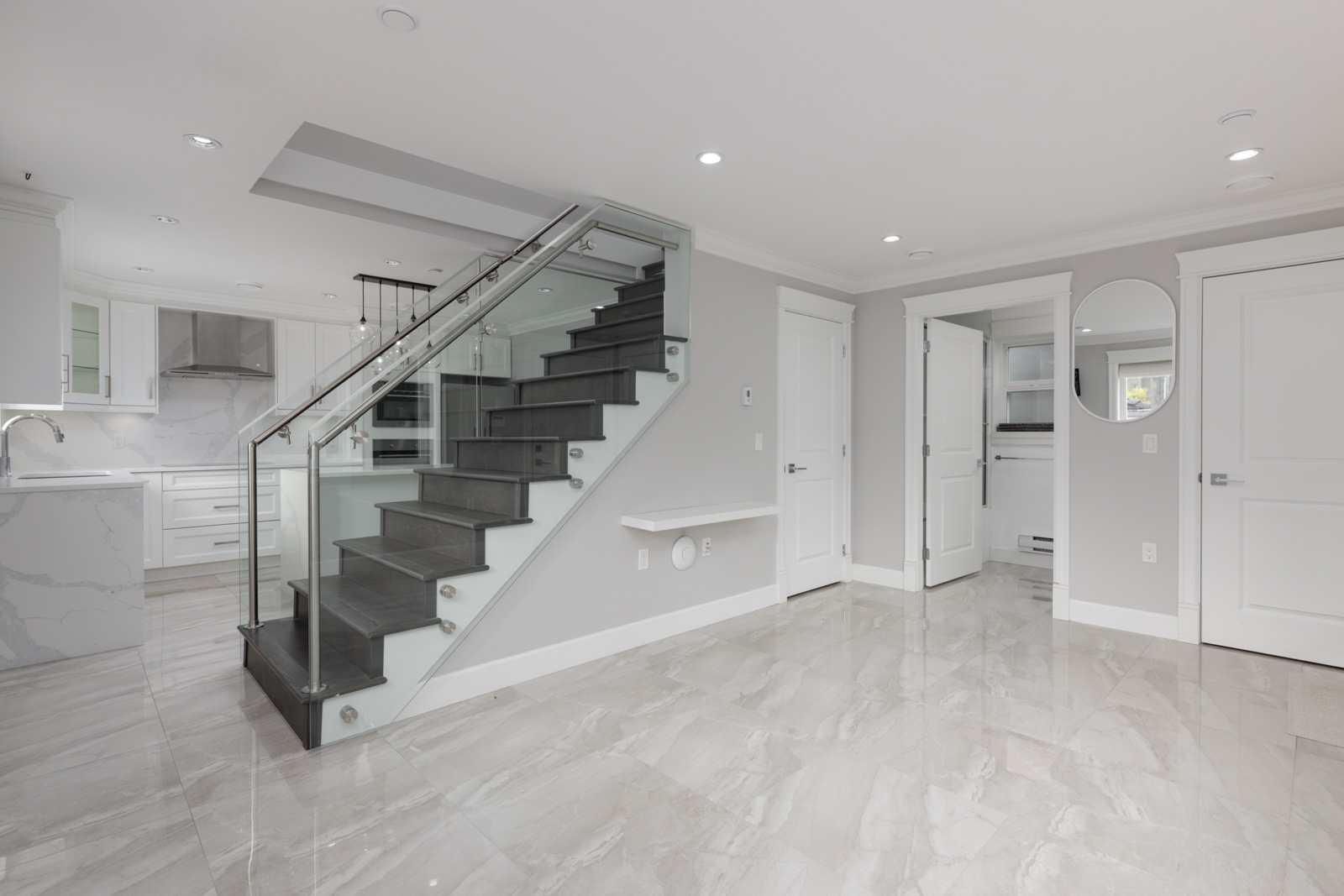 Modern Vancouver interior with gray tile floor, glass staircase, white walls, recessed lighting, and kitchen with white cabinets.