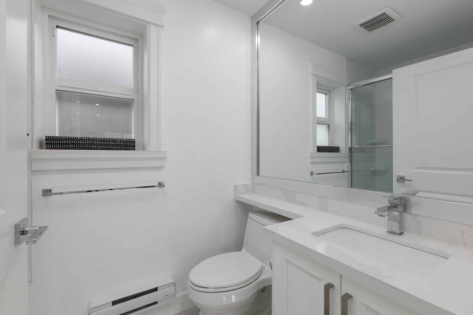 Modern white bathroom with toilet, large mirror, sink, window with black blinds, and towel rack in a Vancouver Falaise Ave home.