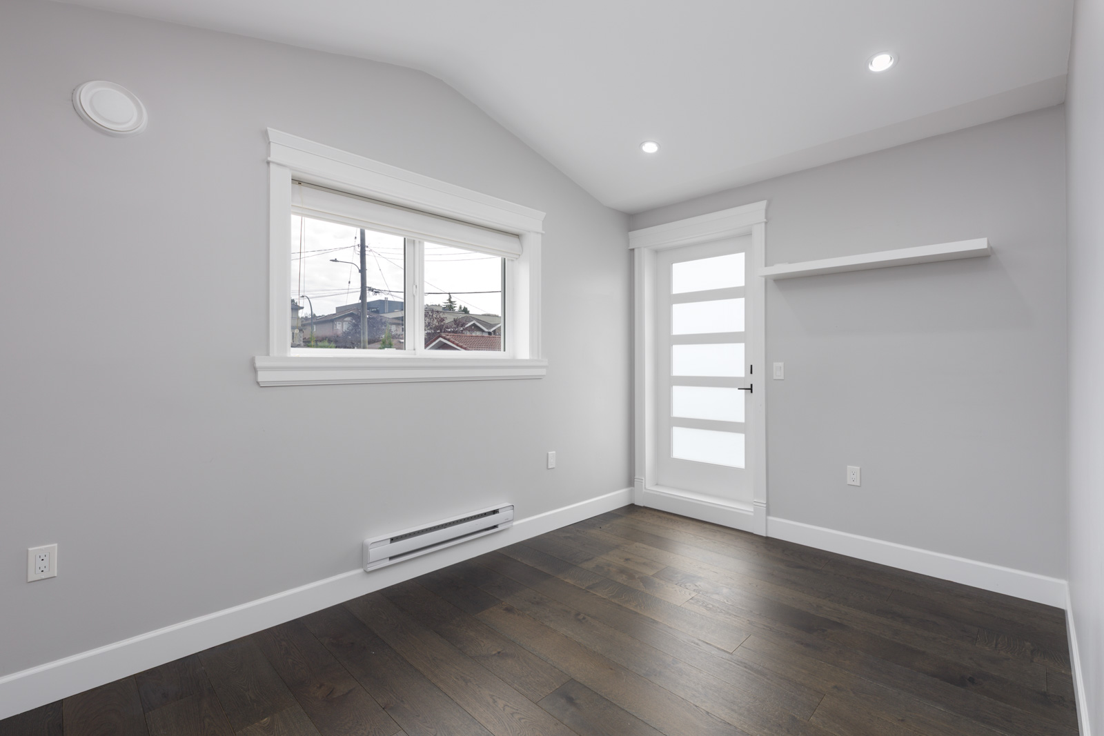 Empty room with light gray walls, dark wood floor, window, white door with frosted glass panels, shelf, recessed lights; Vancouver near Falaise Place.