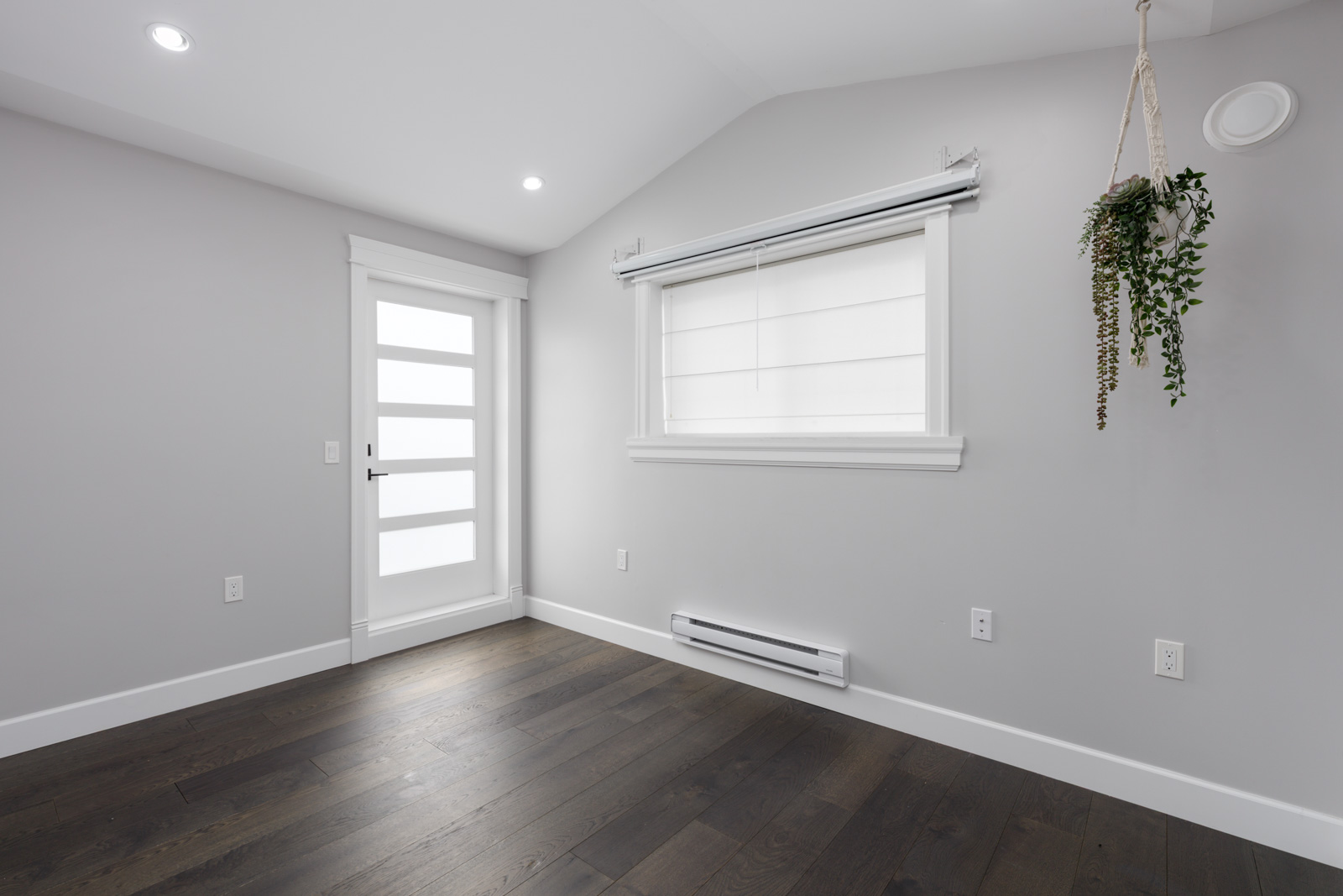 Empty room with light gray walls, dark wood floor, frosted glass door, white-shaded window, and hanging plant in Vancouver.