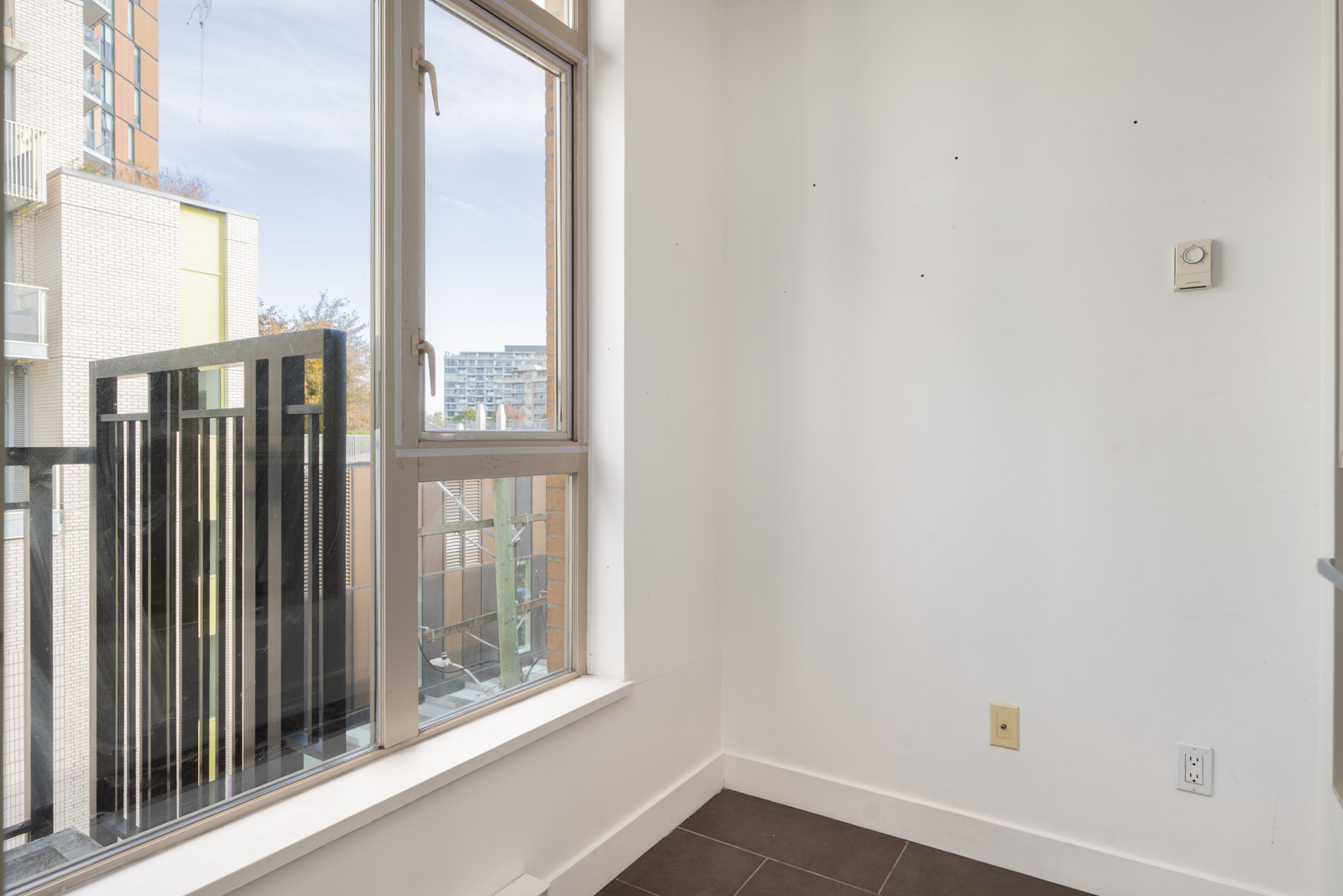 Small empty room with white walls, tiled floor, outlets, and large window facing buildings and balcony on 4th floor in Vancouver.