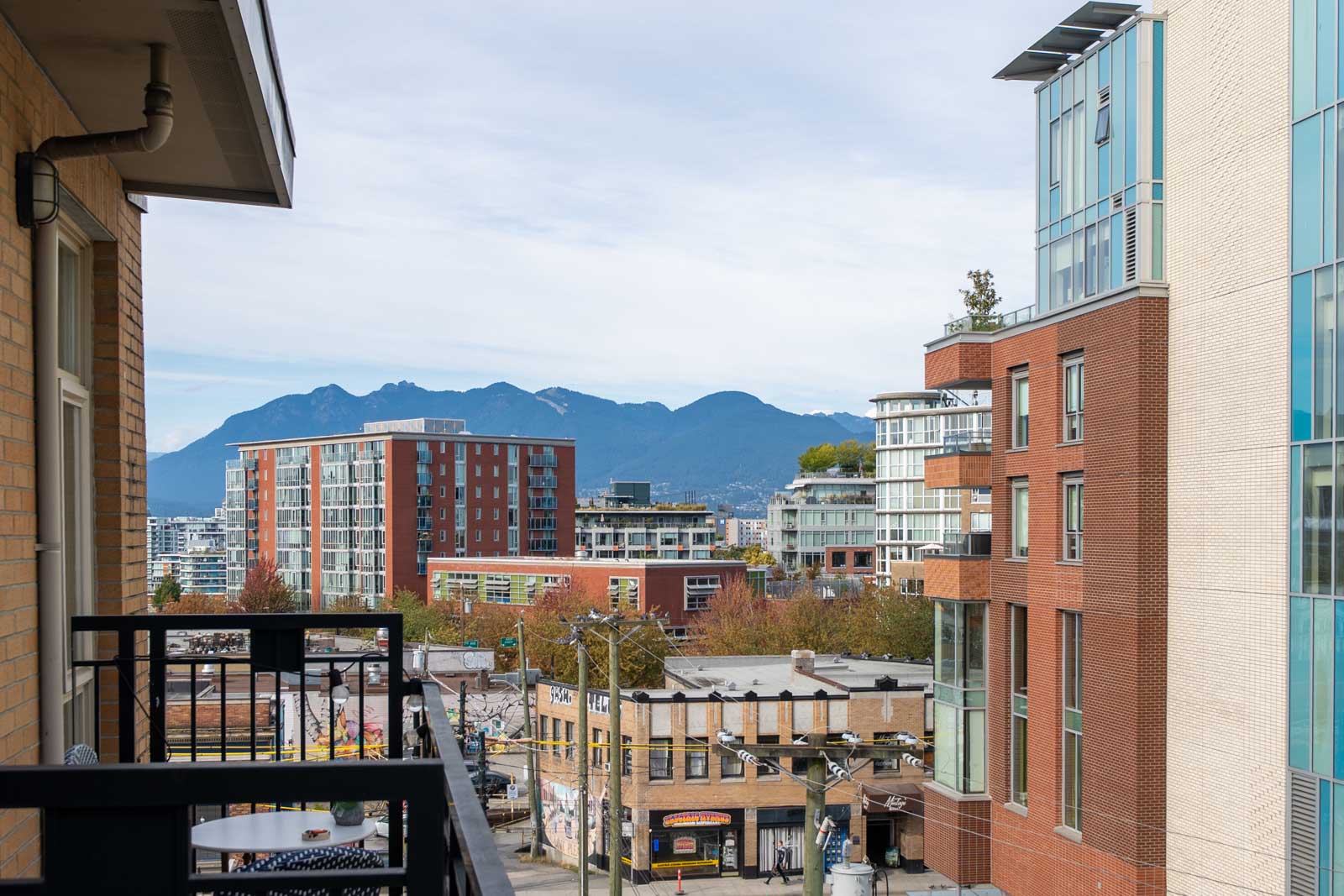 View from a 4th floor balcony at 205 E 10th Ave, Vancouver: city street, modern buildings, trees, and mountains under clouds.