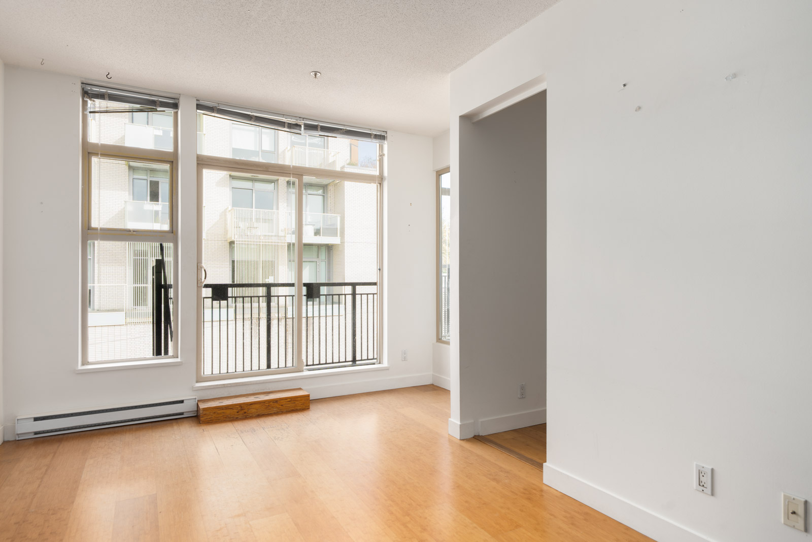 Empty room with large windows, light wood floors, and white walls on the 4th floor in Vancouver, overlooking a balcony and apartments.