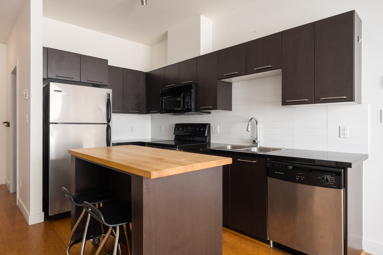 Modern kitchen with dark wood cabinets, stainless appliances, wooden island with two black stools, white tile backsplash; 4th Floor, 205 E 10th Ave, Vancouver.