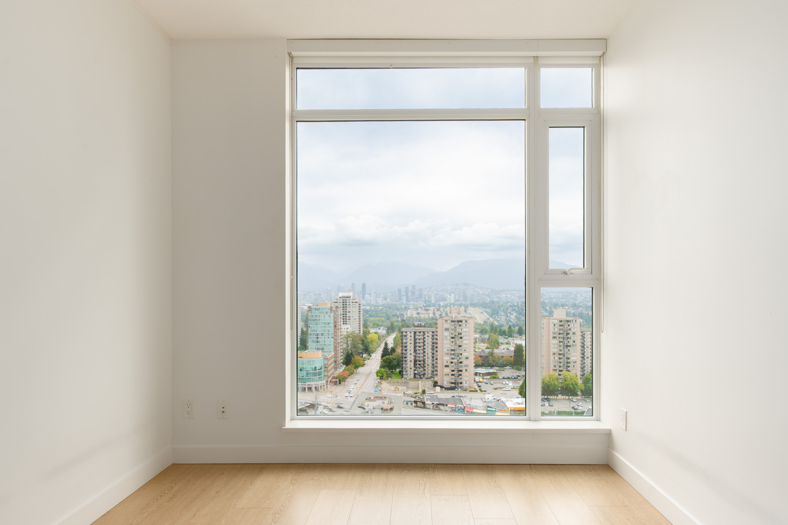 Empty white room with light wood floor and large window overlooking city buildings, trees, and mountains in Burnaby under a cloudy sky.