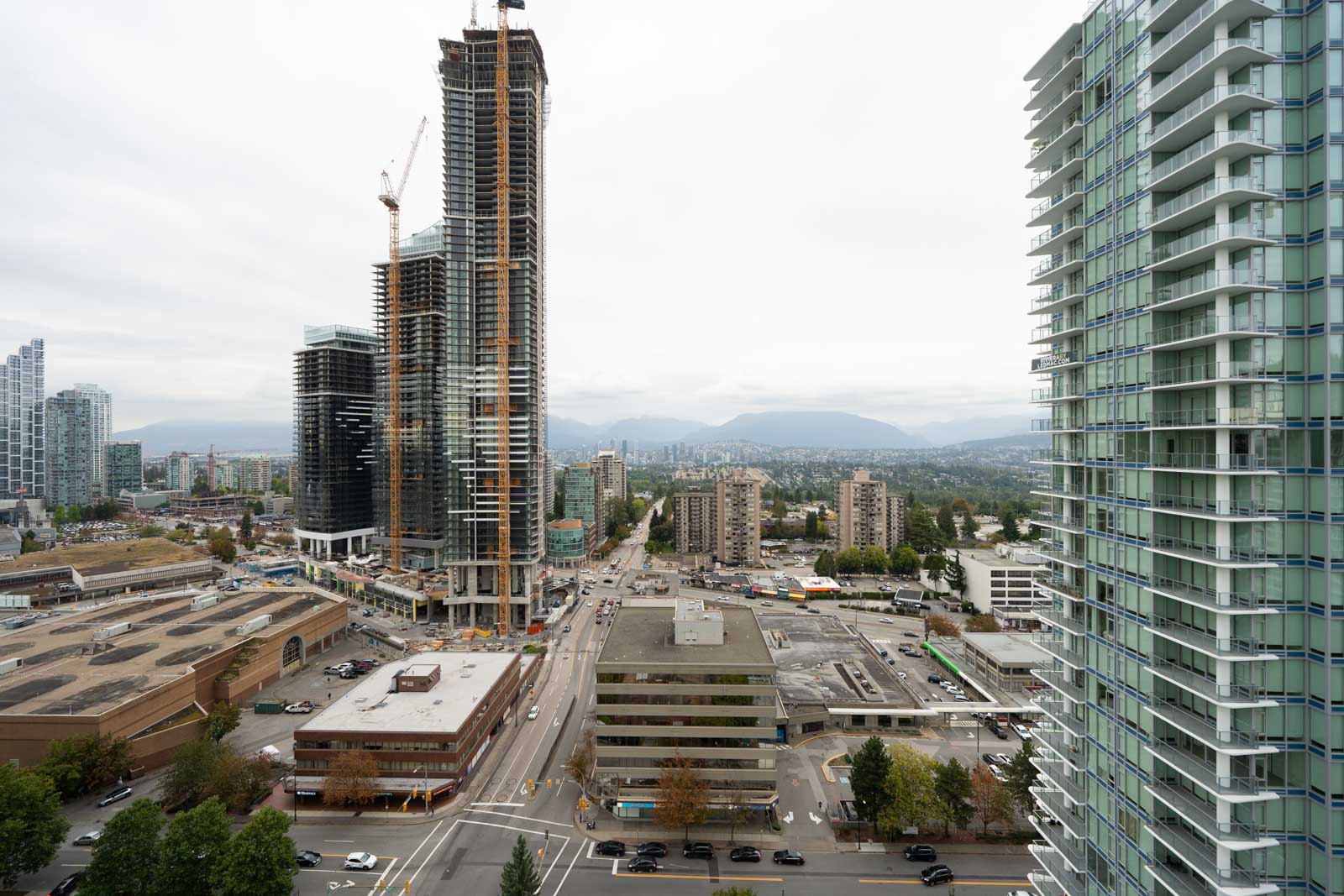 High-rise buildings under construction along Nelson Ave in Burnaby, with parking lots and mountains under a cloudy sky.