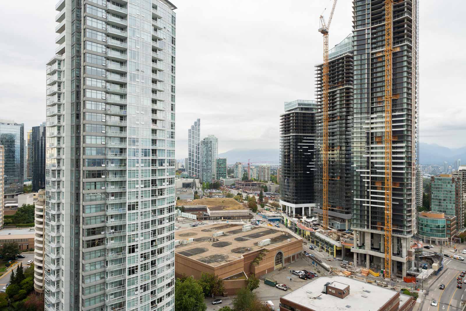 High-rise buildings under construction in urban Burnaby, cranes above Nelson Ave, winding roads, and mountains under a cloudy sky.