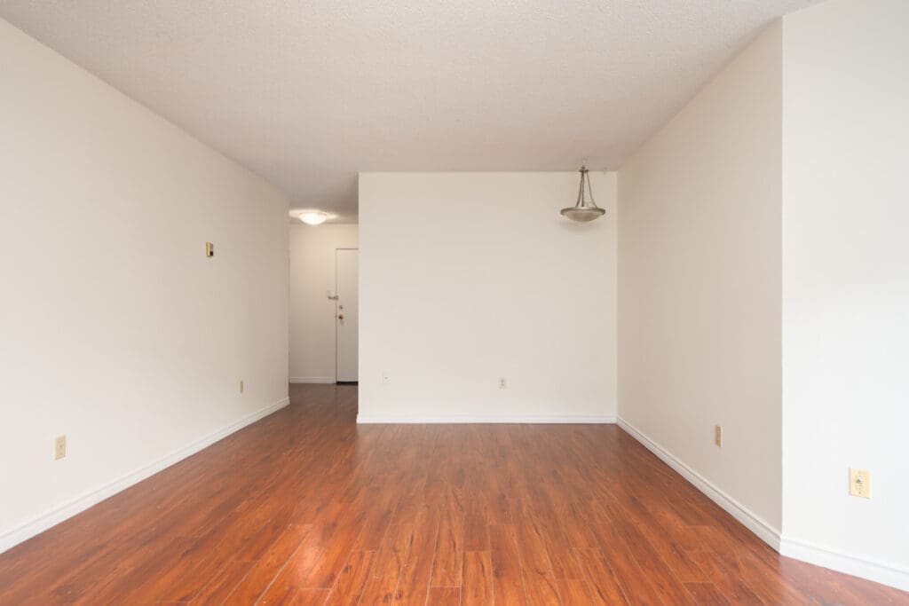 Empty room with white walls, wood floors, small ceiling light, and a hallway to a closed door. Located on Ackroyd Rd, Richmond.