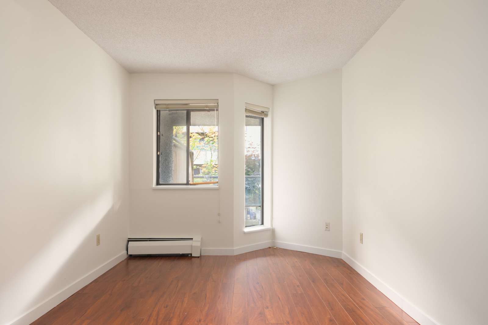 Empty room on Ackroyd Rd, Richmond, with white walls, wood floors, baseboard heater, and two windows bringing in natural light.