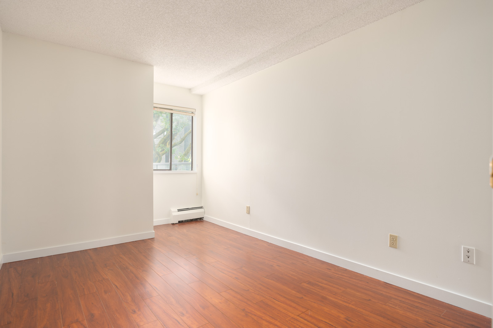 Empty room with light walls, wood laminate floor, a window with blinds, and white baseboard heater in Richmond’s 8460 Ackroyd Rd area.