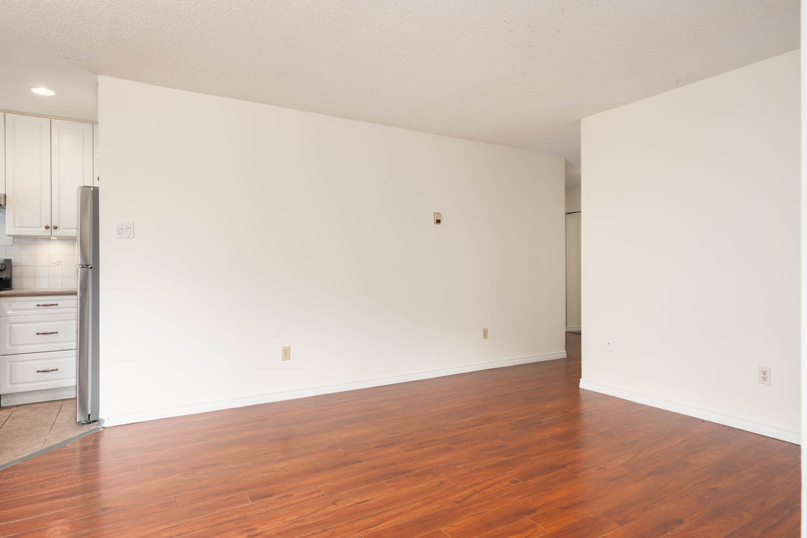Empty room with white walls, wood floors, and a peek of a kitchen with white cabinets and stainless fridge at 8460 Ackroyd Rd, Richmond.