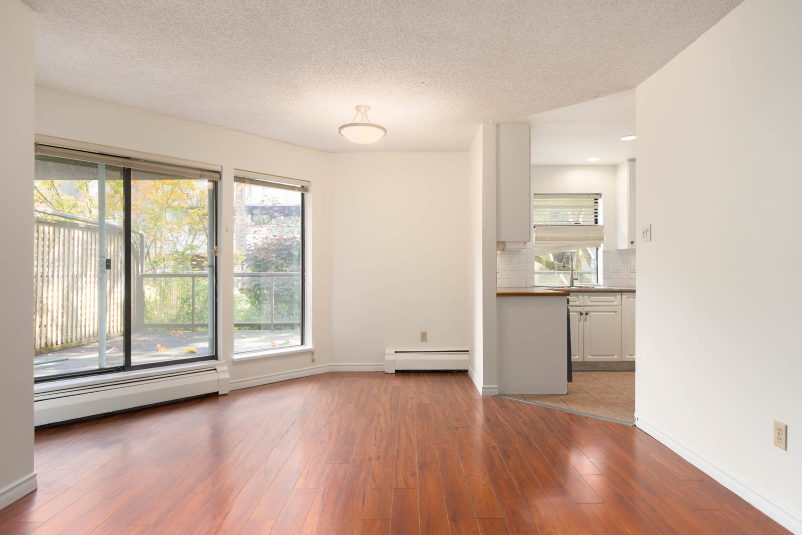 Unfurnished room with wood floors, large windows, sliding glass door to balcony, and partial kitchen view in Richmond’s 8460 Ackroyd Rd.