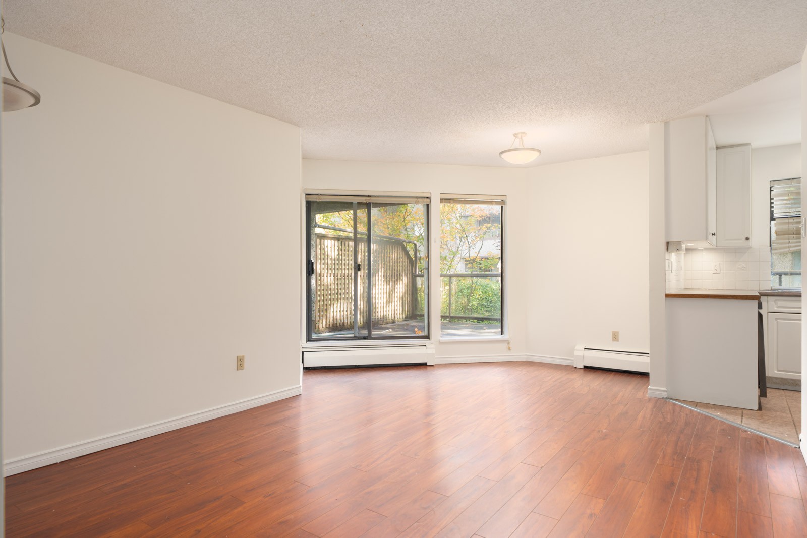 Unfurnished living room with wood floors, large windows, sliding glass door, and glimpse of white kitchen in Richmond.