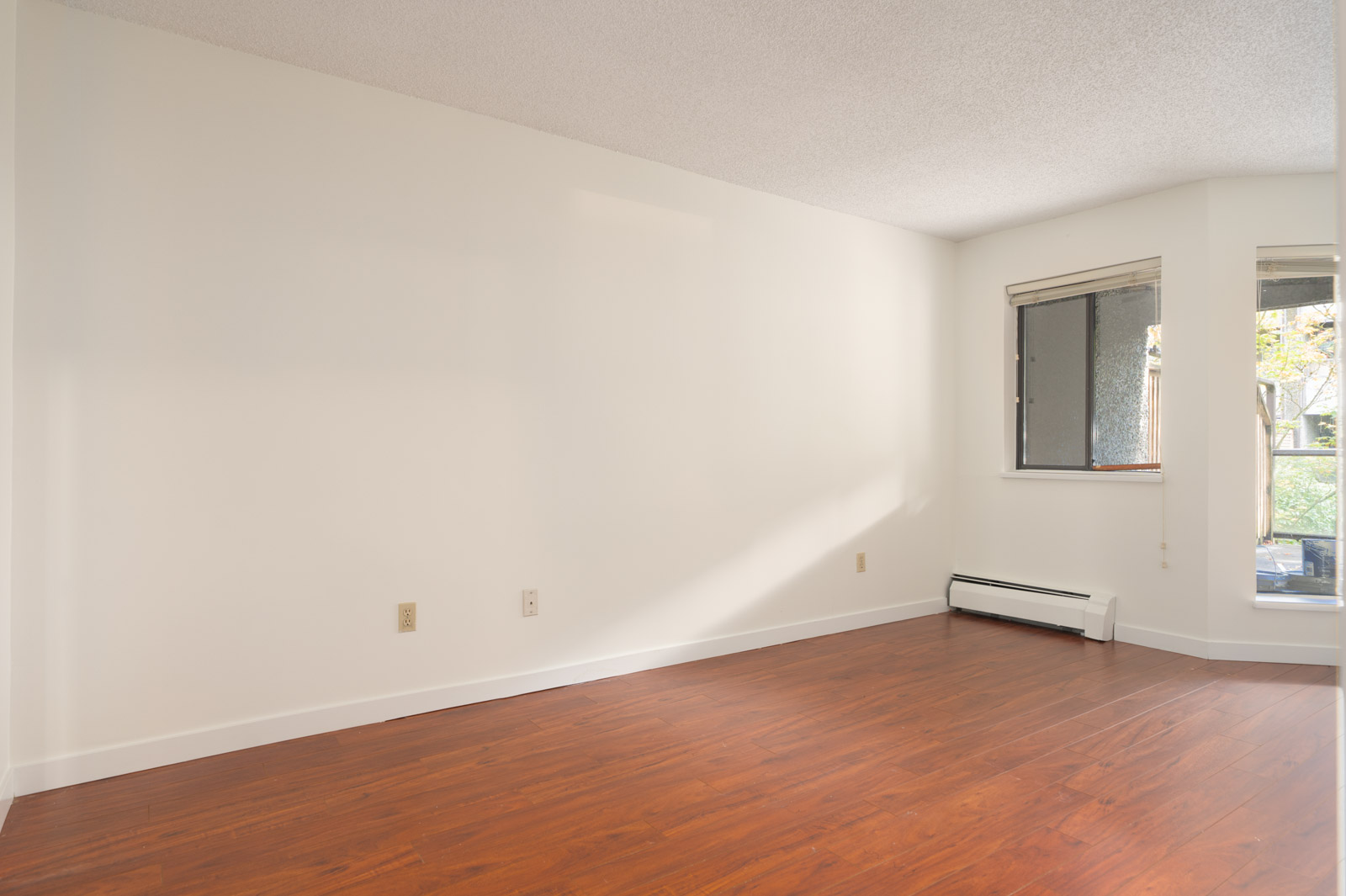 Empty room with white walls, wood floor, window with sunlight, baseboard heater, and outlet on 1st floor at 8460 Ackroyd Rd, Richmond.