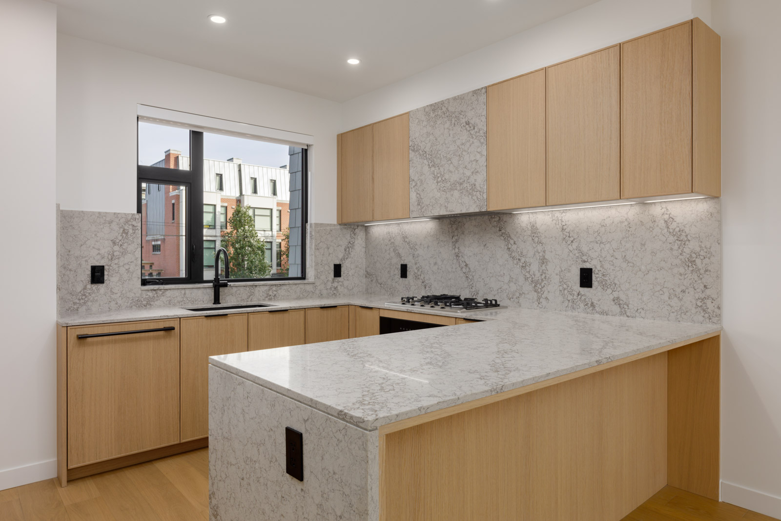 U-shaped kitchen in a Vancouver townhouse featuring light wood cabinets, gray-veined stone counters, and a city window.