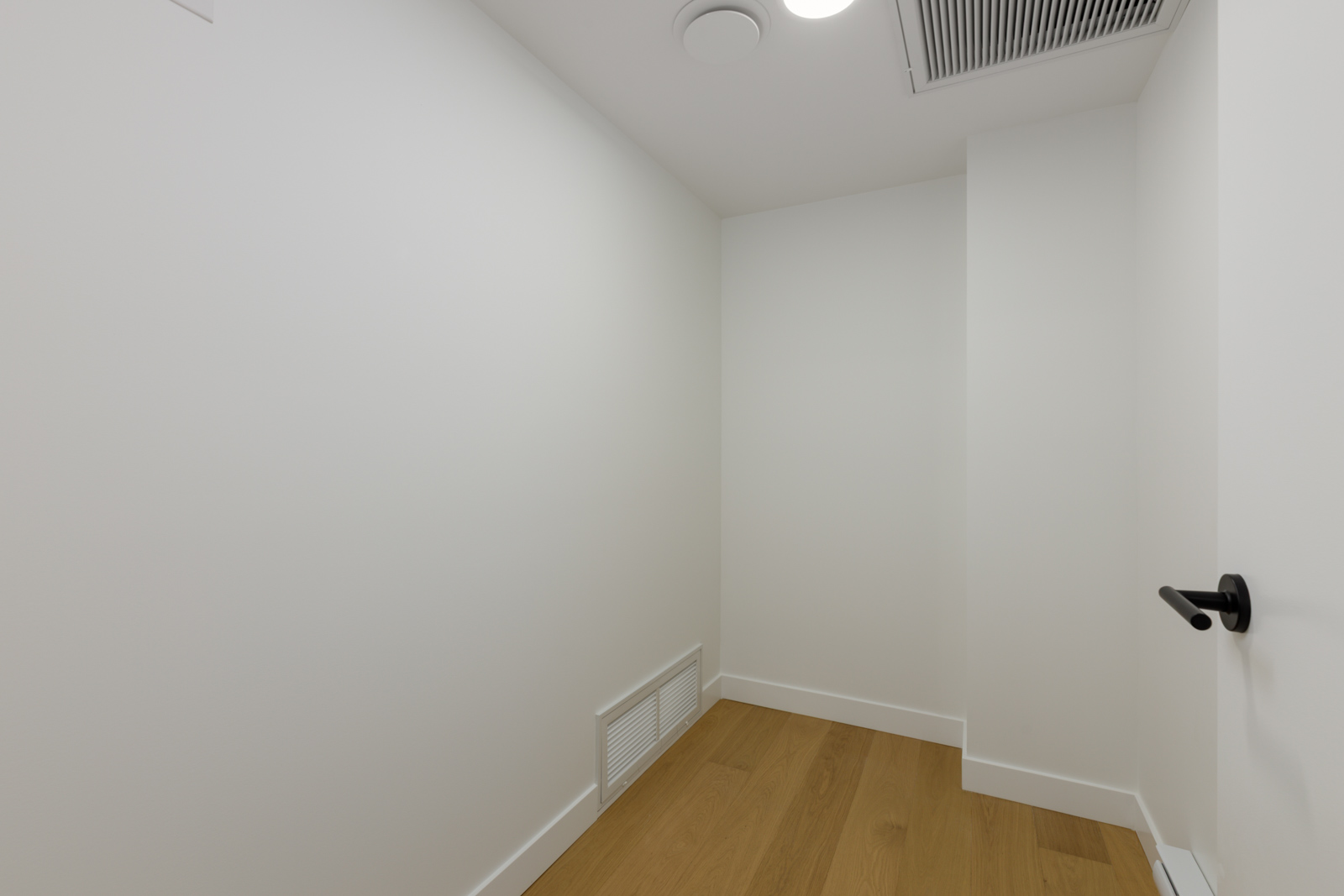 Small, empty white room at 5433 Oak Street with wooden floors, a wall vent, and a black door handle on the right.