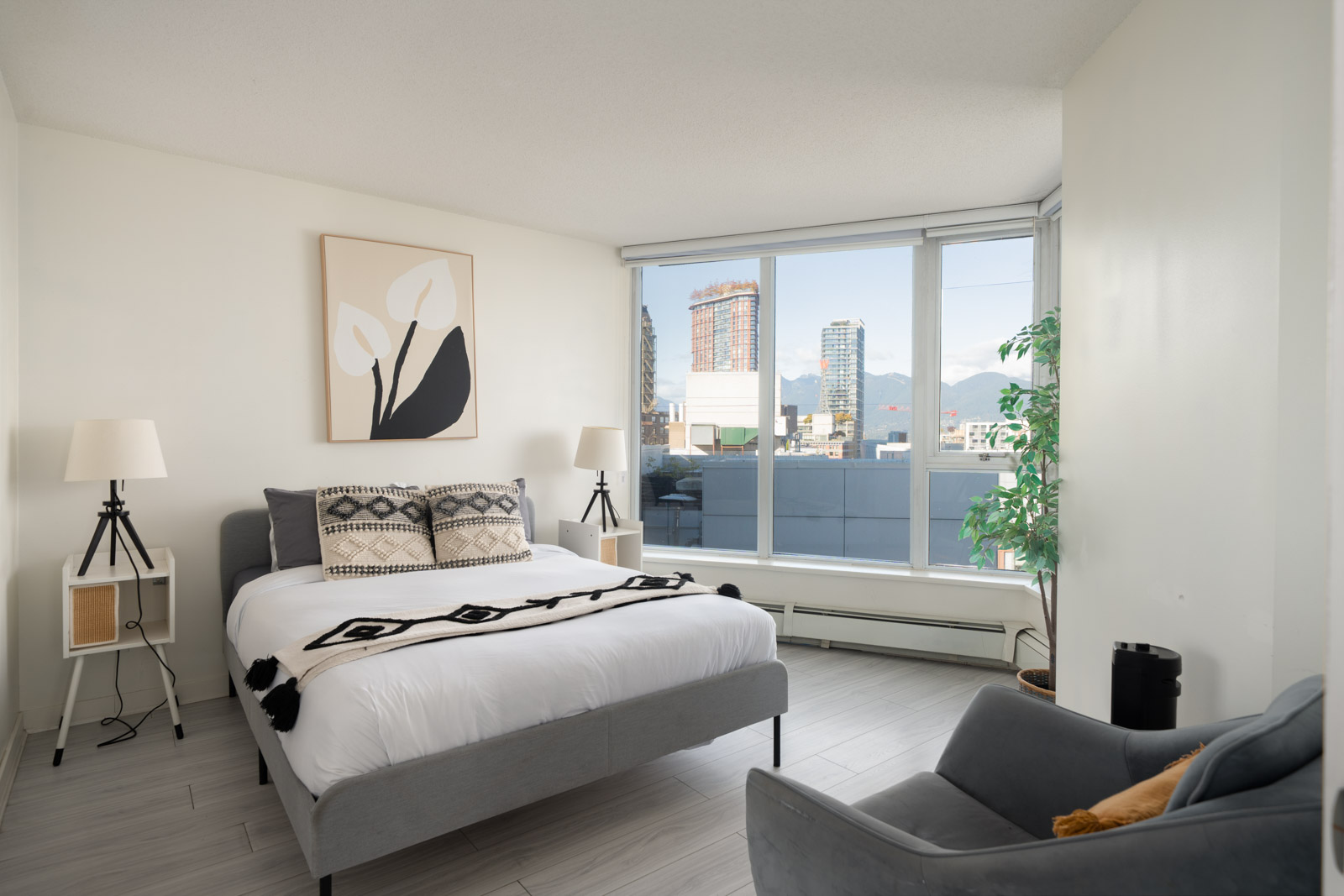 Modern bedroom at 689 Abbott St featuring a gray bed, patterned pillows, and floor-to-ceiling views of the Vancouver skyline.