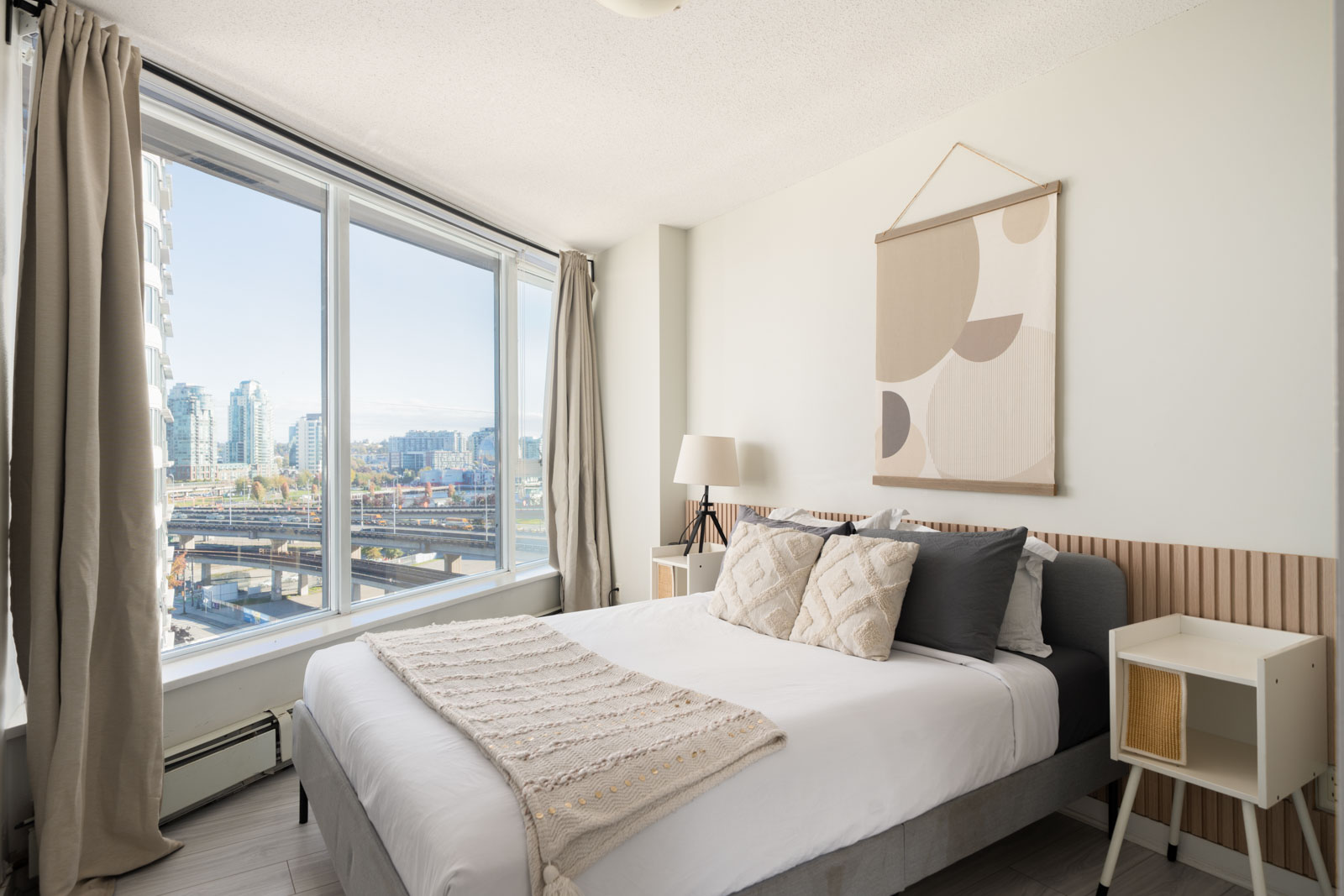 A bedroom with a bed and nightstands, featuring a large window with a 10th-floor view of the city skyline.