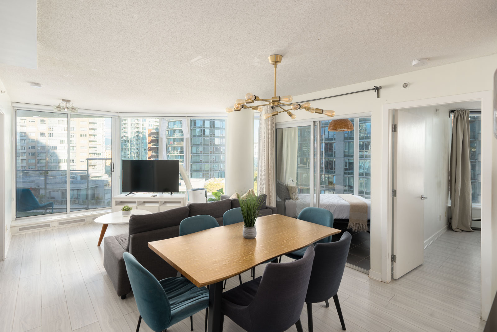 Modern dining area with a wood table and blue chairs, living room with sofa and TV, overlooking Vancouver city high-rises.