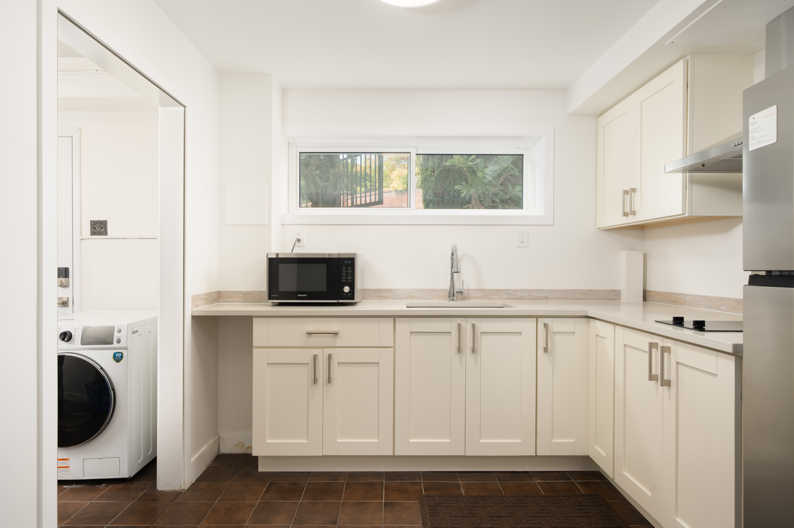 Vancouver basement kitchen with cream cabinets, a countertop microwave, a stainless steel sink, and an adjacent laundry area.