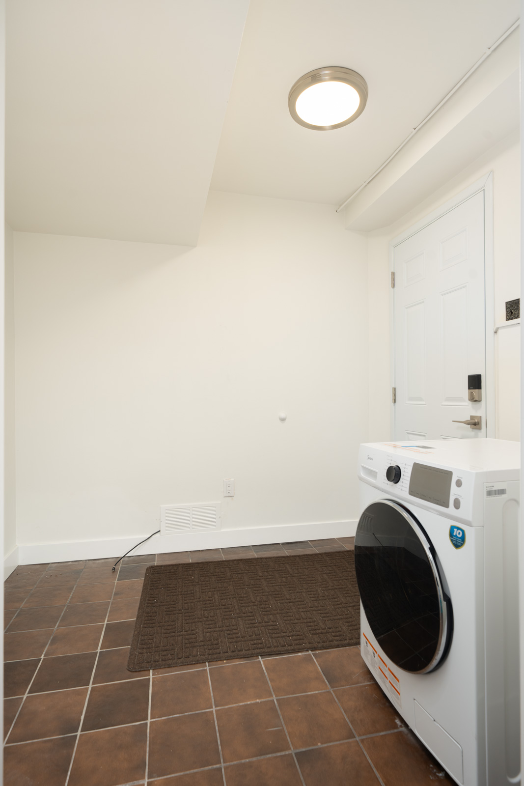 White front-loading washing machine in a Vancouver basement with brown tiled floors and a white door.