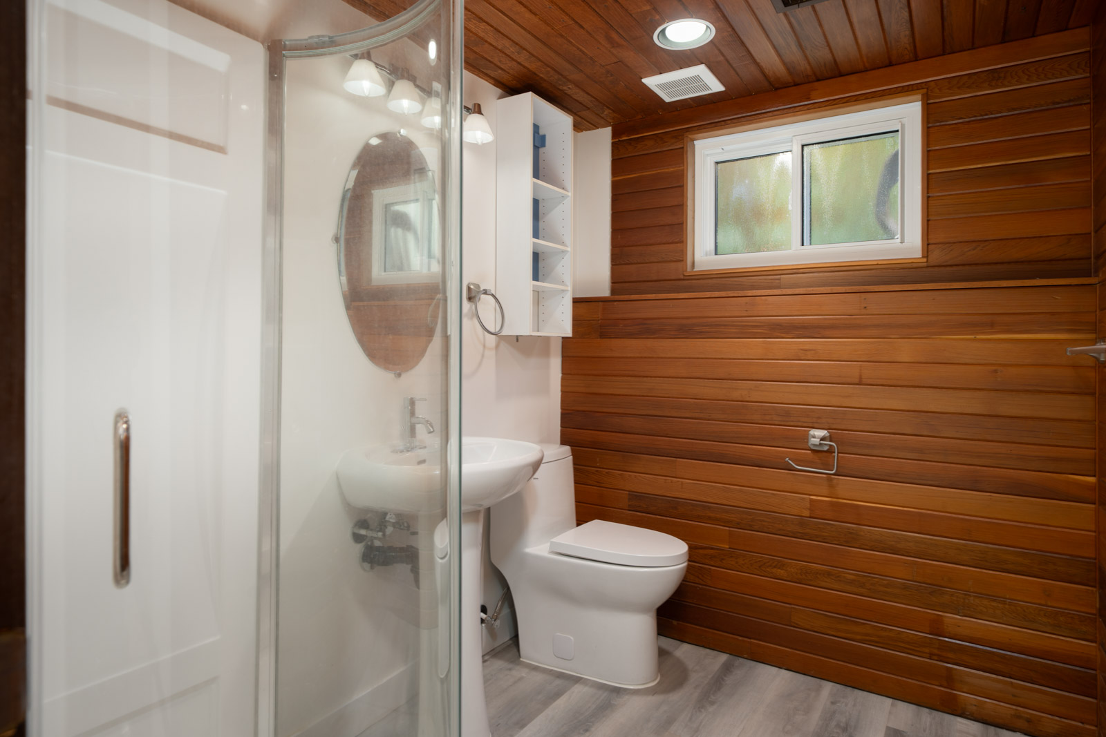 Vancouver basement bathroom featuring wood-paneled walls, a pedestal sink, toilet, and a glass-enclosed shower.