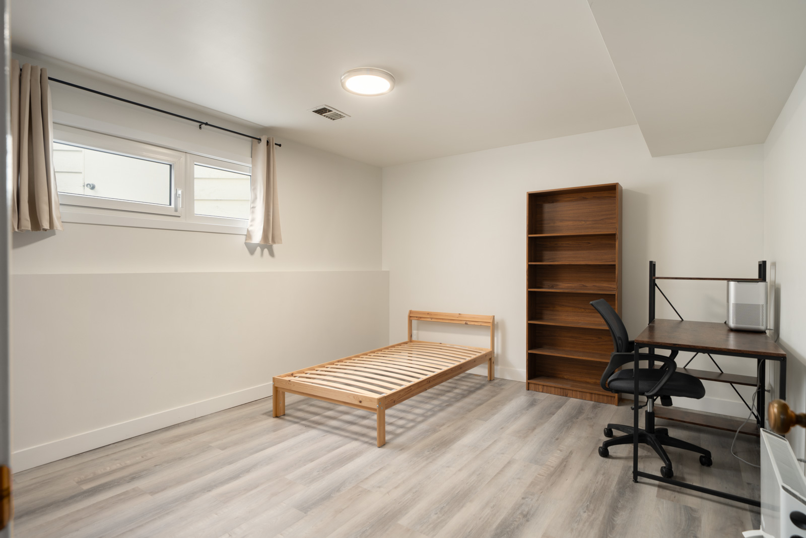 Unfurnished Vancouver basement bedroom with a wooden bed frame, tall bookshelf, desk, and office chair.
