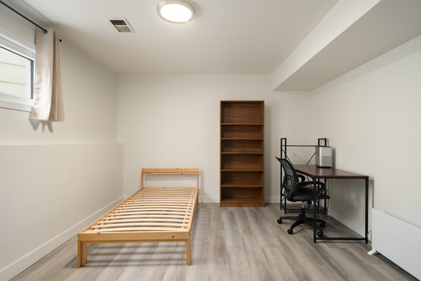 Furnished room in a Haggart Street, Vancouver basement with a wooden bed, tall bookshelf, desk, and office chair.