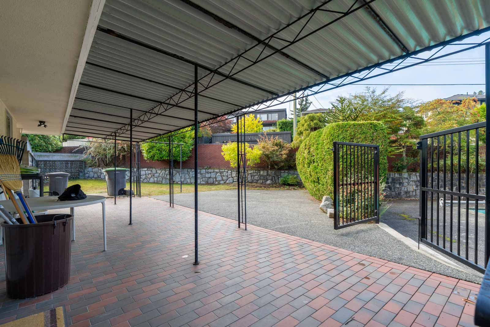 A paved patio at a Vancouver basement, sheltered by a metal carport, with a gate opening to a driveway and backyard.