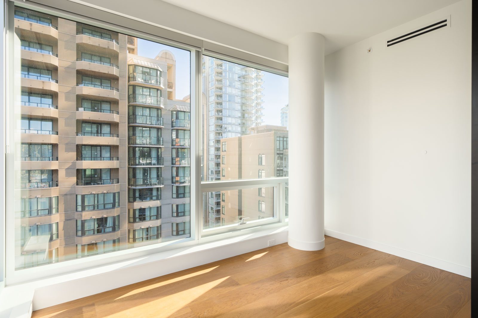 Empty 14th-floor room in Vancouver featuring hardwood floors, a large white column, and windows overlooking the city.