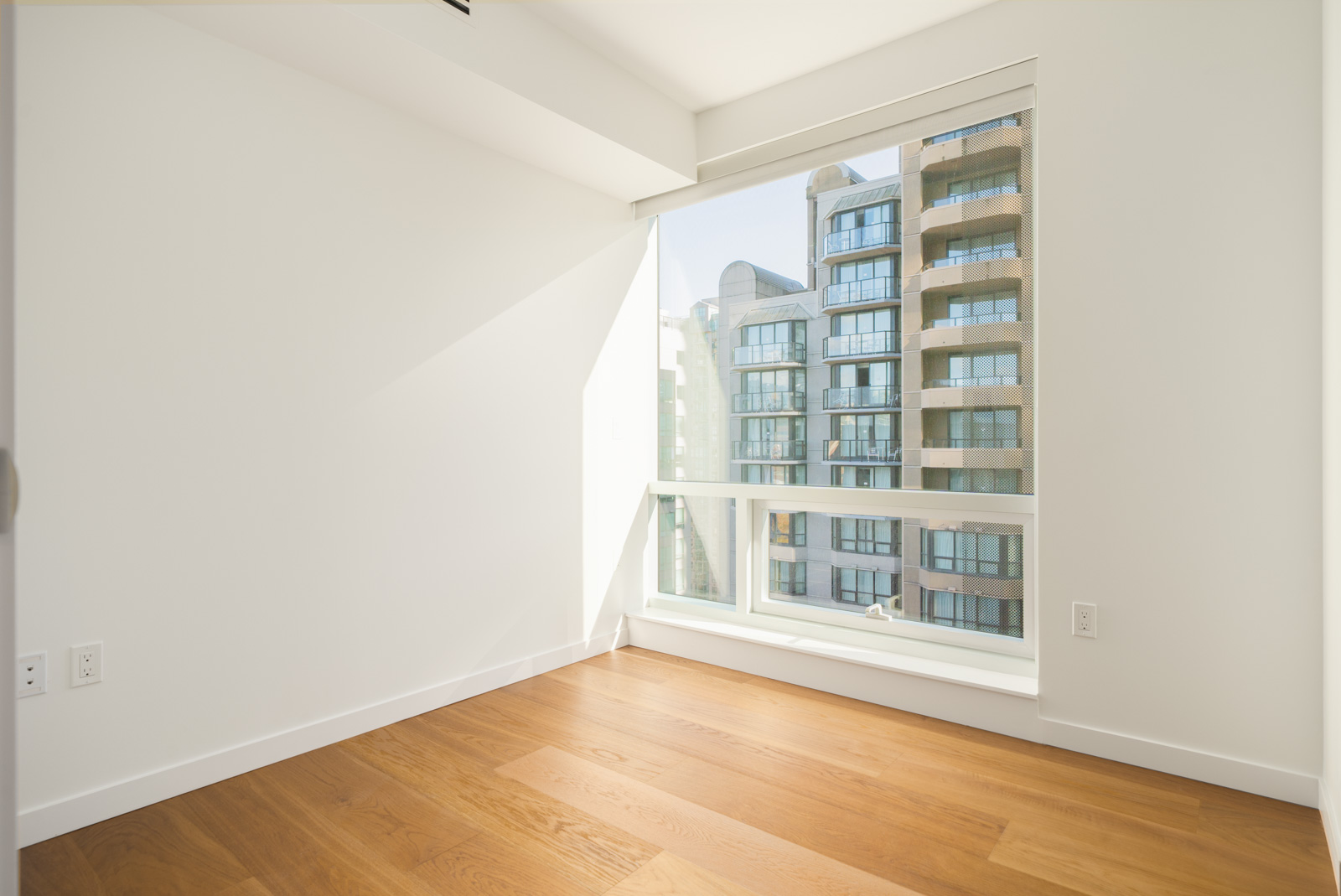 Empty room with hardwood floors and a large window overlooking Vancouver apartment buildings at 1277 Hornby St.