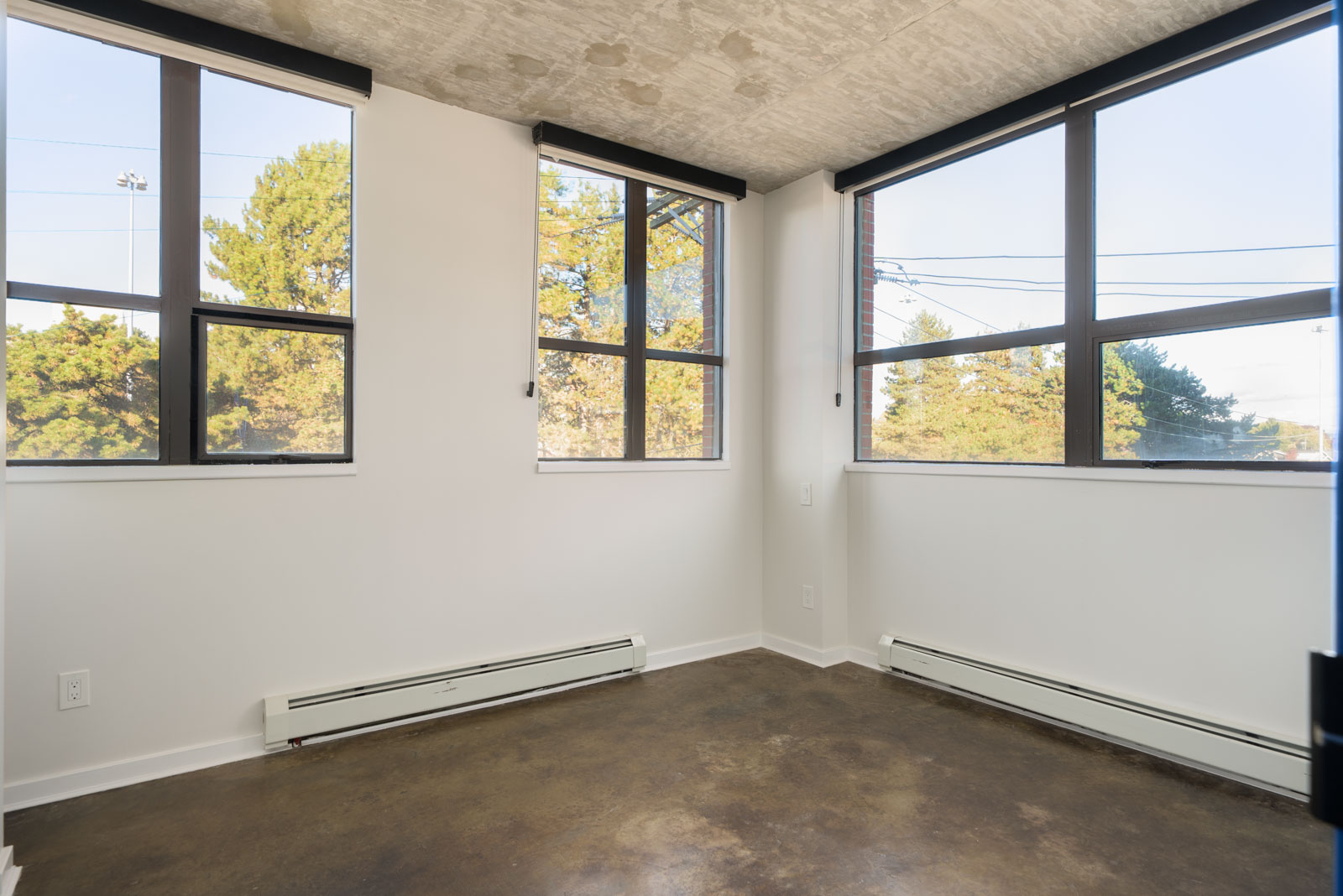 Empty corner room at 919 Station Street with white walls, concrete floors, large windows, and baseboard heaters.