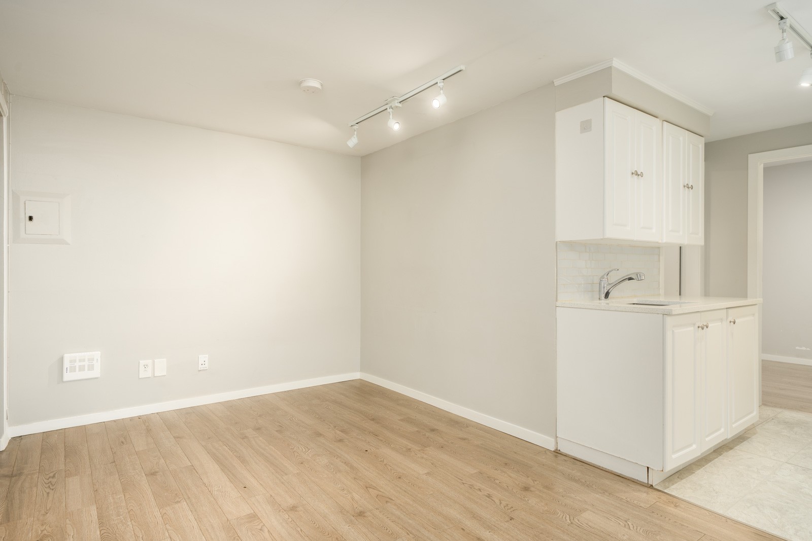 Bright Vancouver rental room with light wood flooring, a plain wall, and a kitchenette with white cabinets.