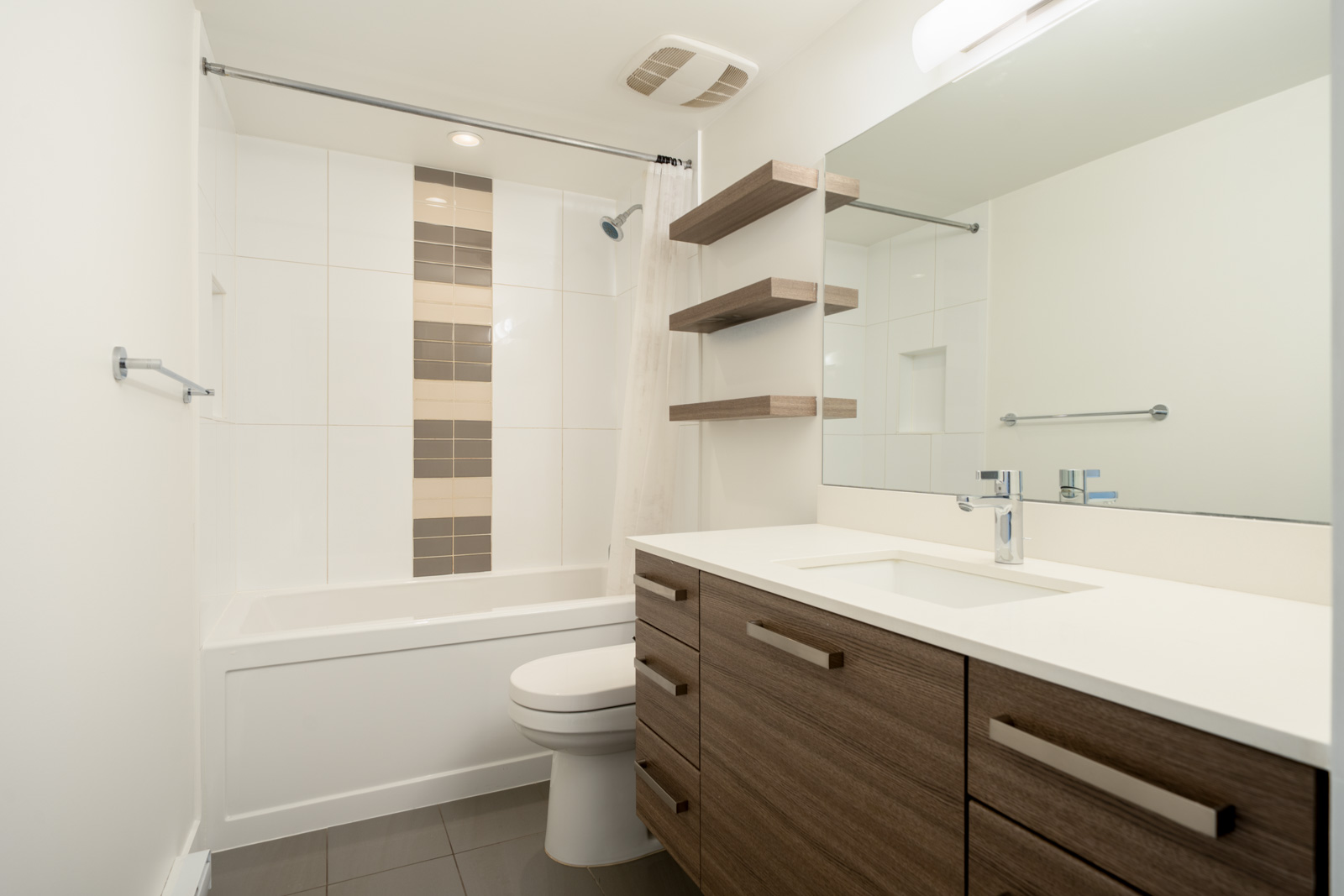 Modern bathroom at 2858 W 4th Ave with a wood vanity, white countertop, toilet, bathtub, tiled accent wall, and shelves.