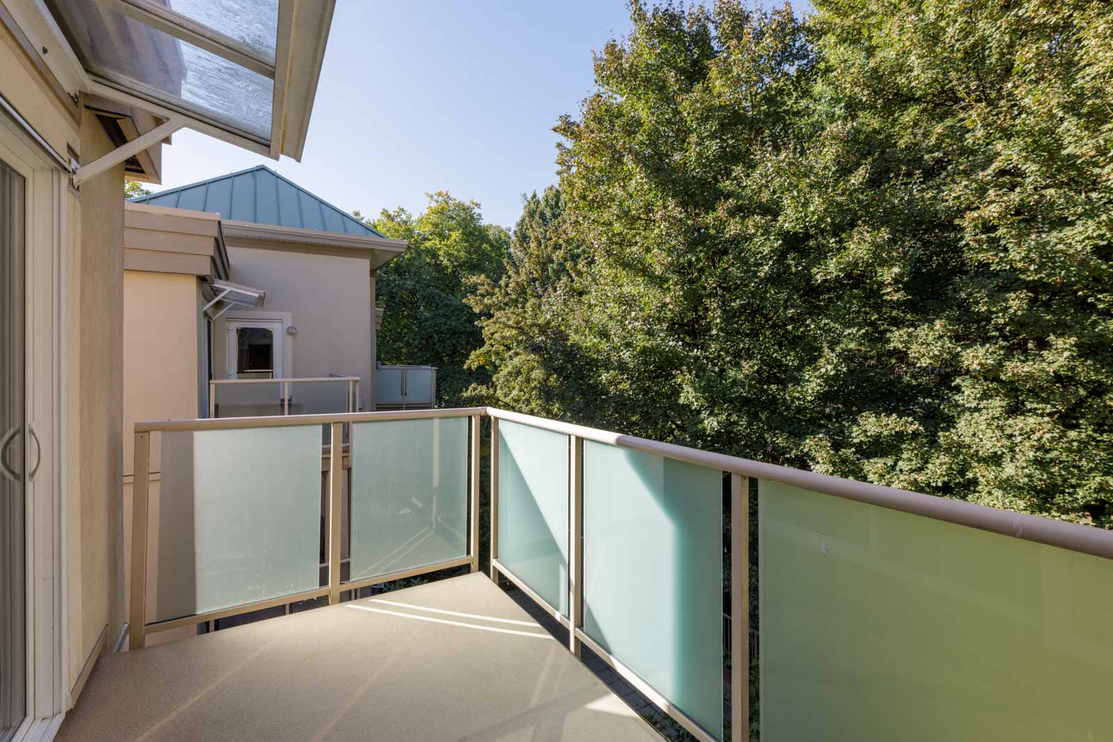 Small 3rd-floor balcony with frosted glass railings overlooks trees and beige buildings on Welcher Ave, Port Coquitlam, under clear sky.