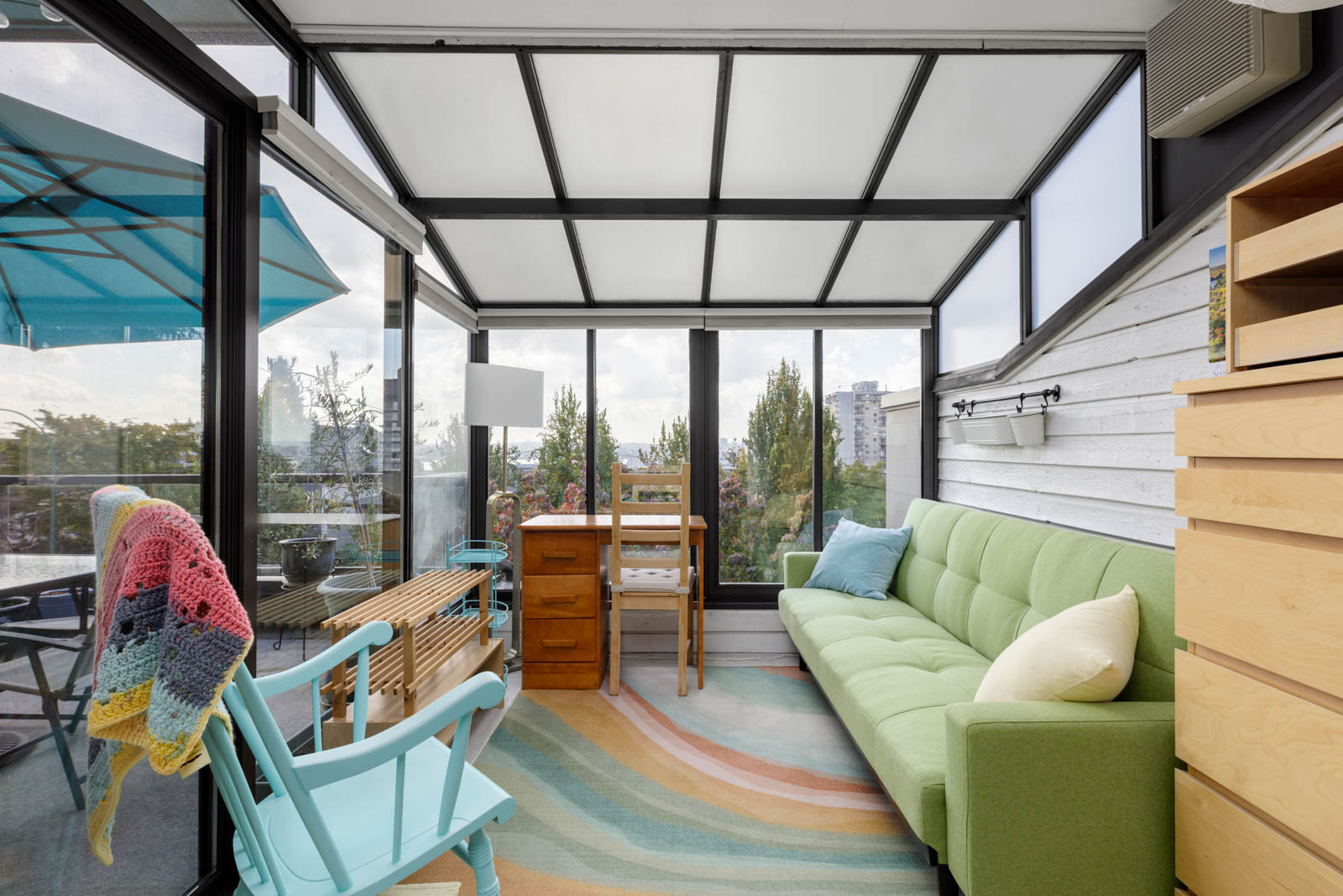 Sunroom in a 4th St E townhouse with glass walls, green sofa, wood desk, blue rocker, rug, and city view through windows.