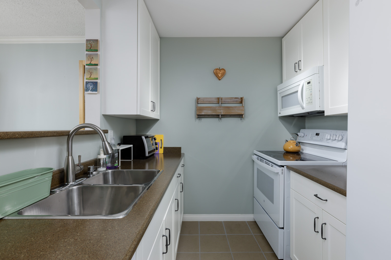 Small North Vancouver kitchen with white cabinets, brown countertops, double sink, green dish rack, and heart decor on green wall.