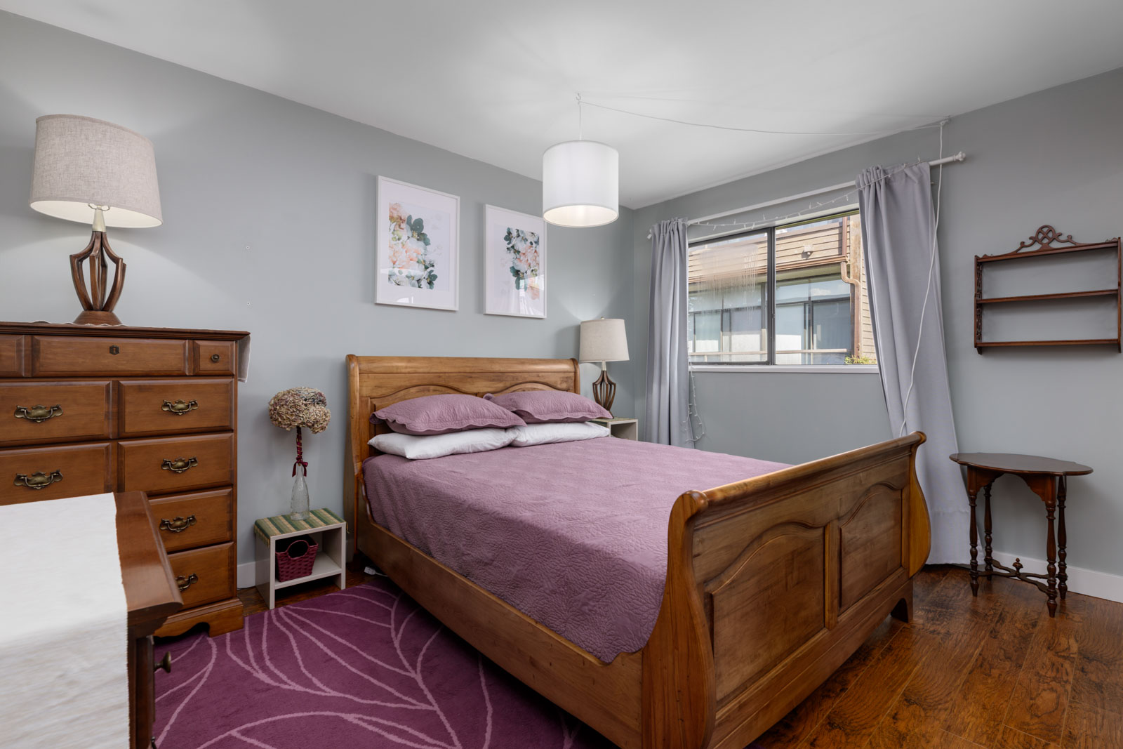 Bedroom in a North Vancouver townhouse with a wooden bed, purple bedding, floral art, gray curtains, and hardwood floors.