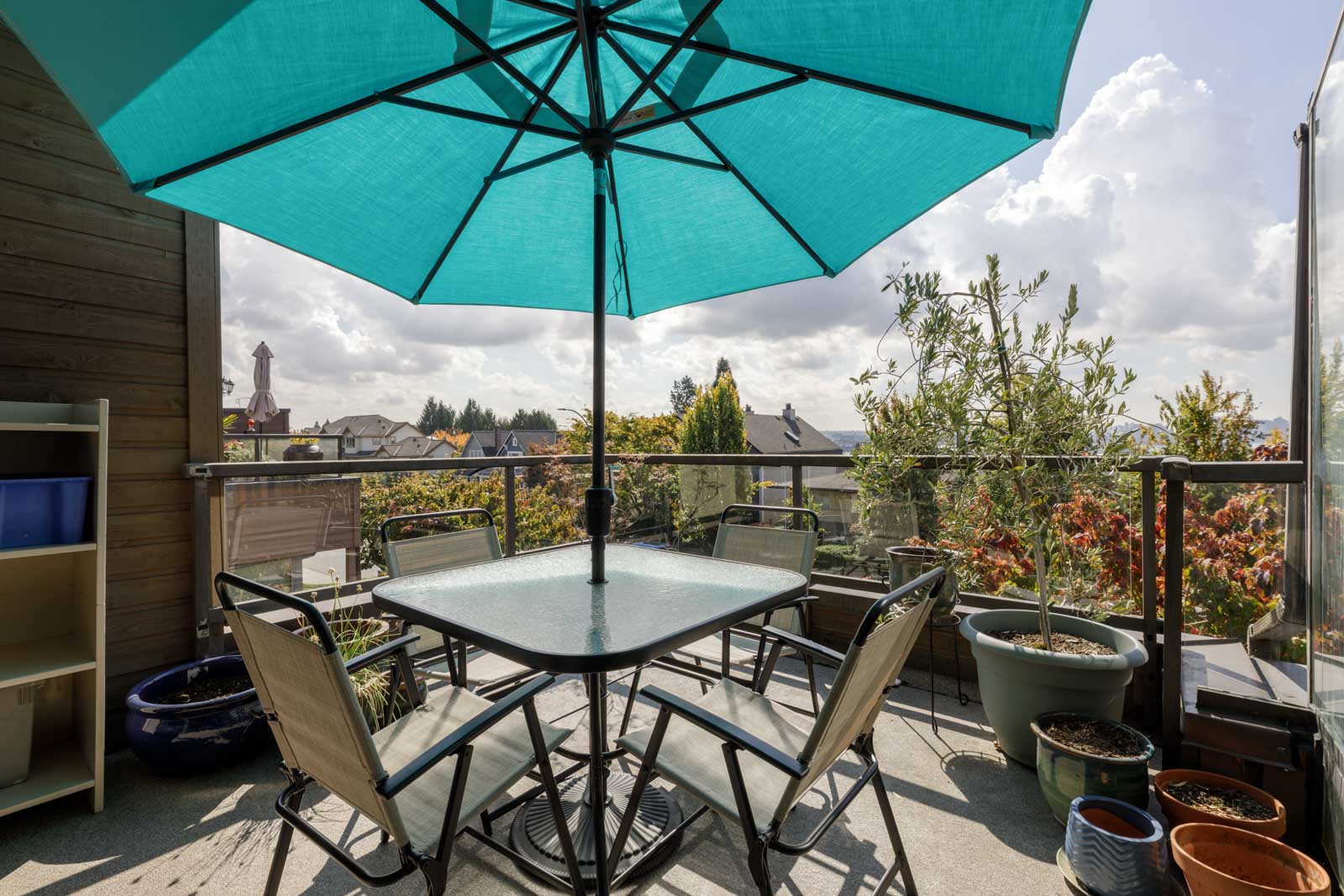 Sunny outdoor patio at 220 4th St E with glass table, six chairs, teal umbrella, and surrounding potted plants and trees.