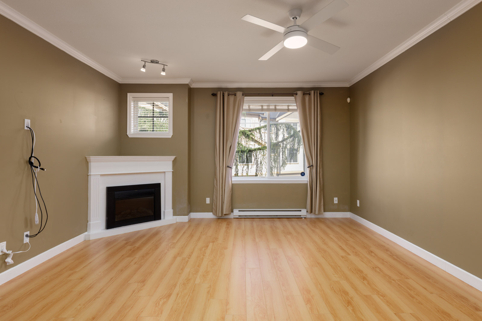 Empty living room in a Surrey townhouse featuring light wood floors, tan walls, a white fireplace, and a curtained window.
