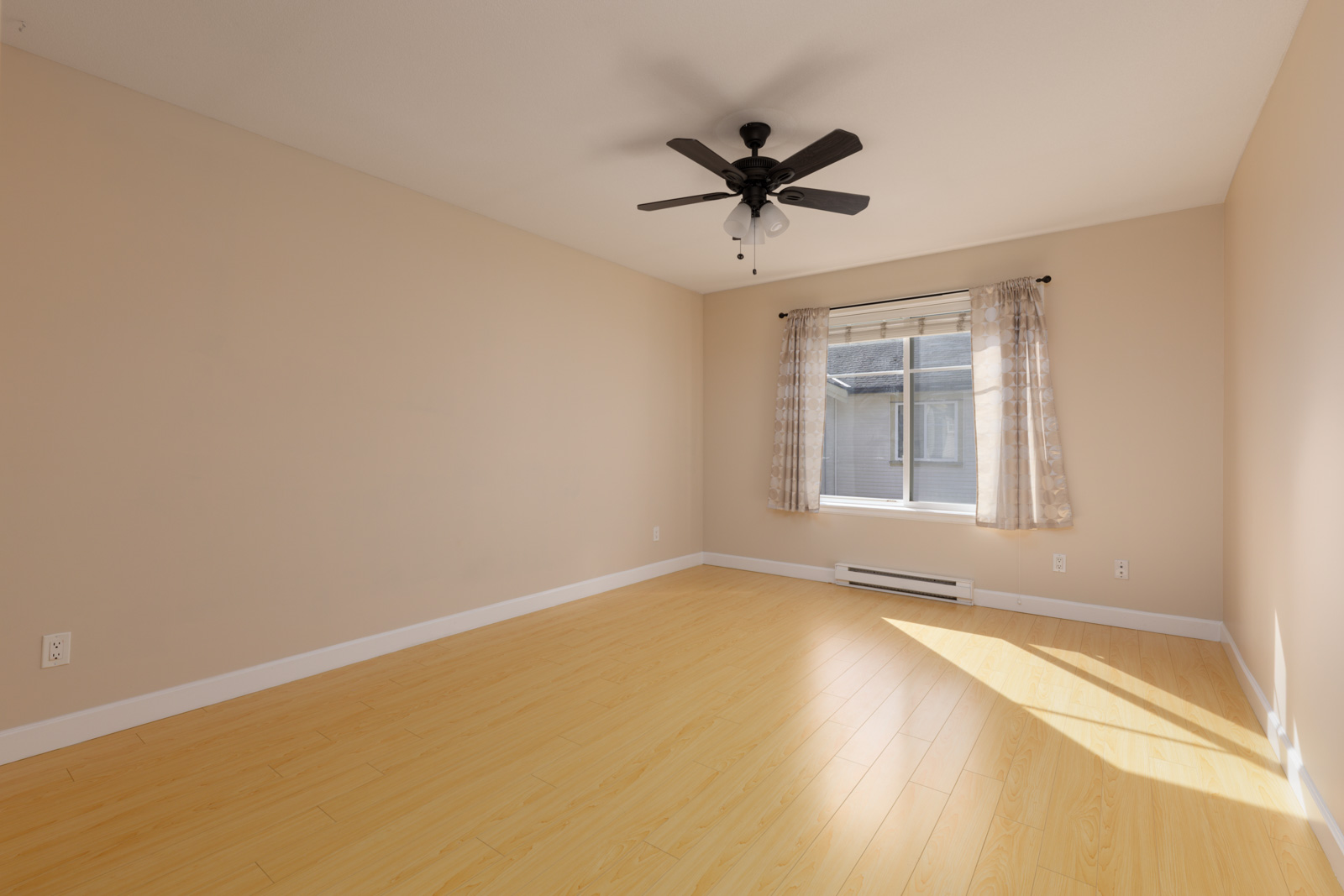 Empty Surrey townhouse room with beige walls, light wood flooring, a ceiling fan, and a window with curtains.