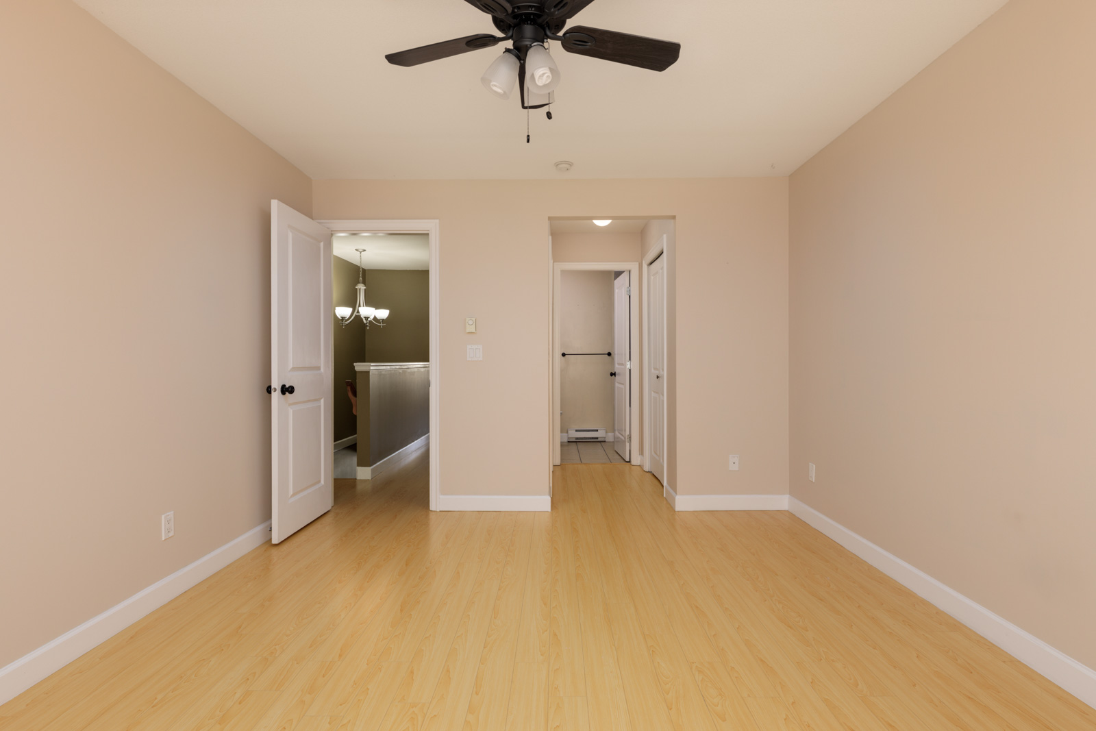 An empty room in a Surrey townhouse featuring light wood floors, a ceiling fan, and open doorways to a hall and bathroom.