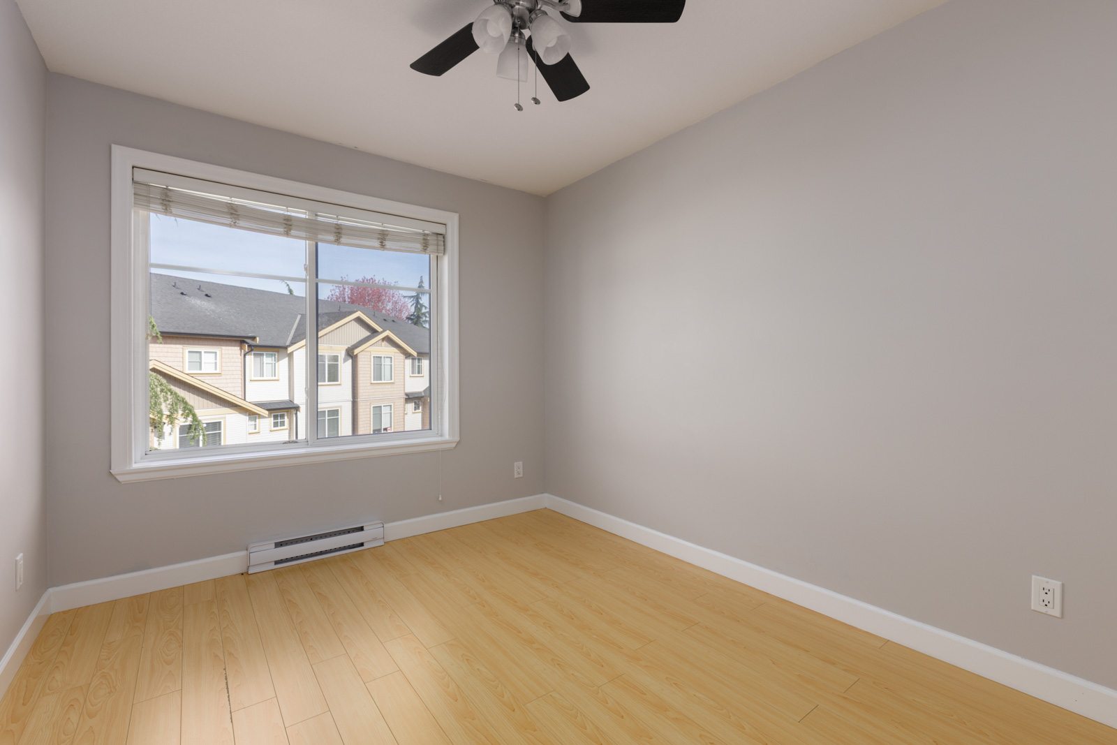 Empty room with light wood floors, gray walls, a ceiling fan, and a window view of Surrey townhouses.