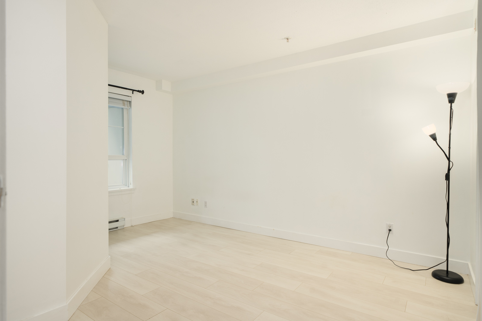 An empty room in a Richmond townhouse featuring white walls, light flooring, a single window, and a black floor lamp.